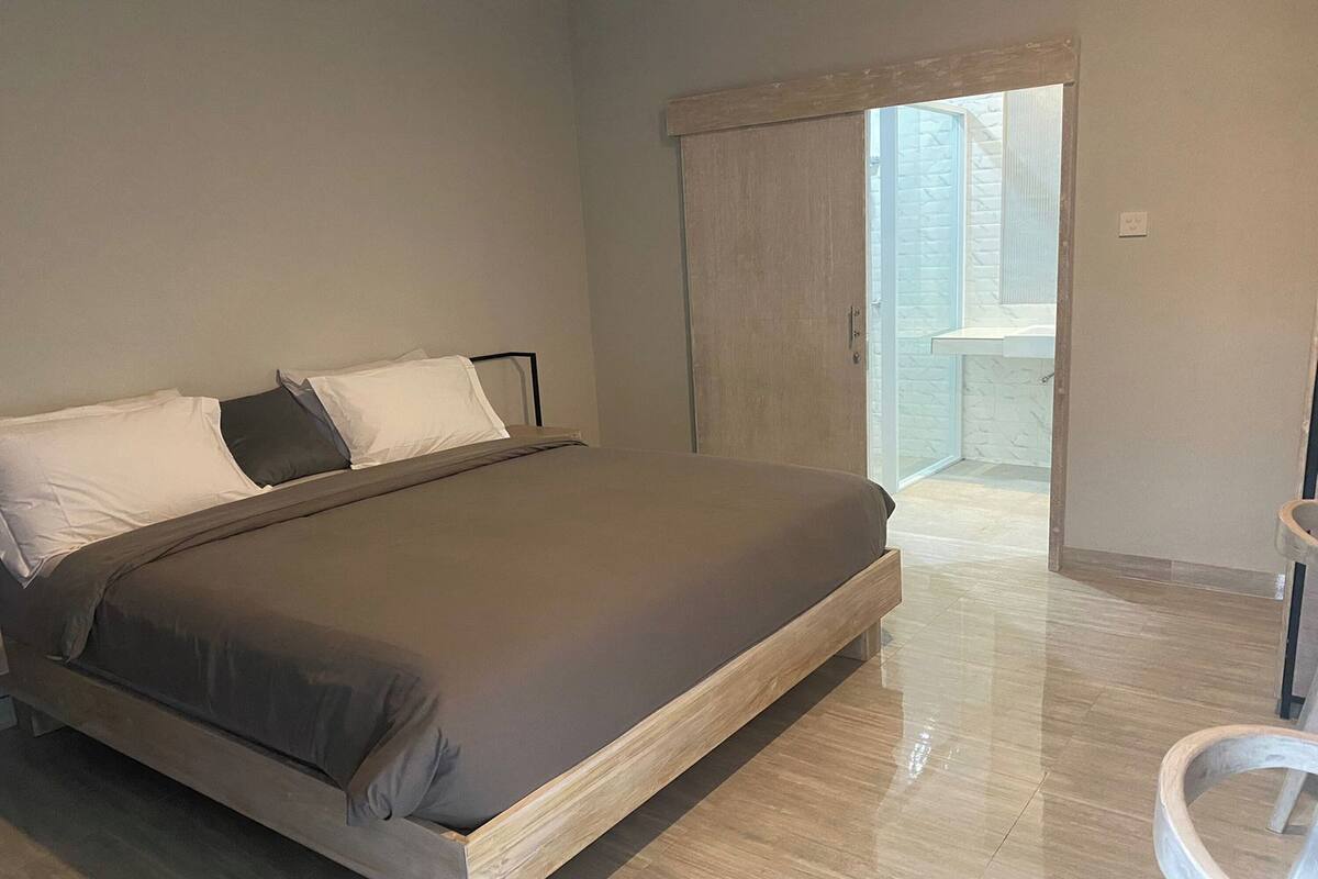 A bedroom with a large bed, a wooden floor, a sliding door leading to a balcony, and a small bedside table.