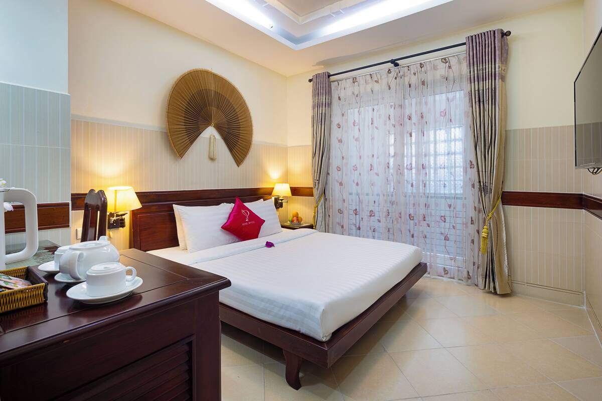 A hotel room with a bed, bedside tables, lamps, a wall fan, and a window with curtains.
