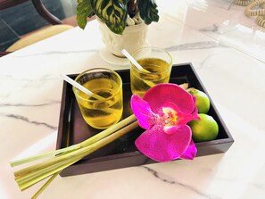 Una bandeja con dos vasos de líquido amarillo, una orquídea rosa y limones verdes sobre una mesa de mármol.