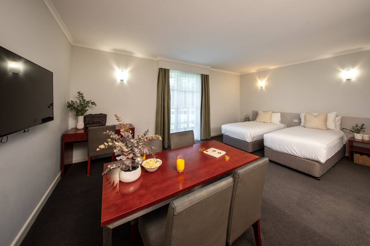 Habitación de hotel con una mesa de comedor de madera, dos camas, un televisor y una ventana con cortinas.
