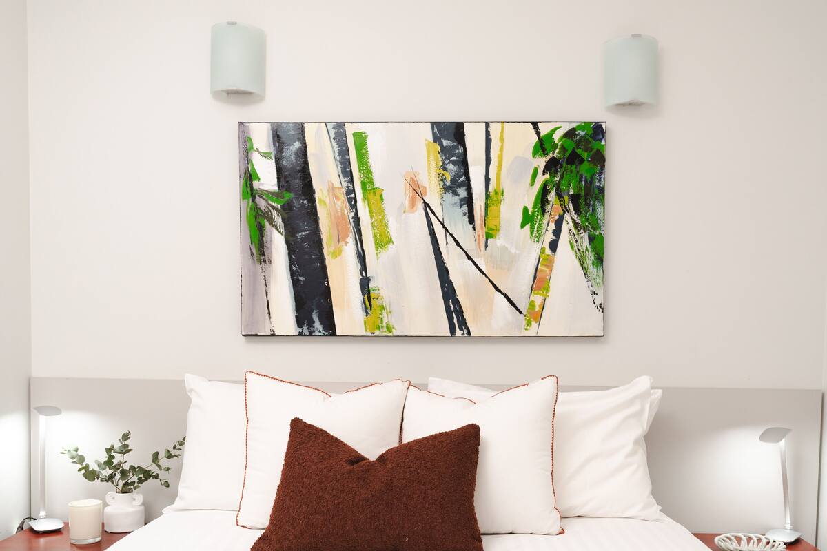 Habitación de hotel con cama, almohadas blancas, un cojín decorativo marrón, un cuadro abstracto en la pared, mesitas de noche con lámparas y un florero con hojas verdes.