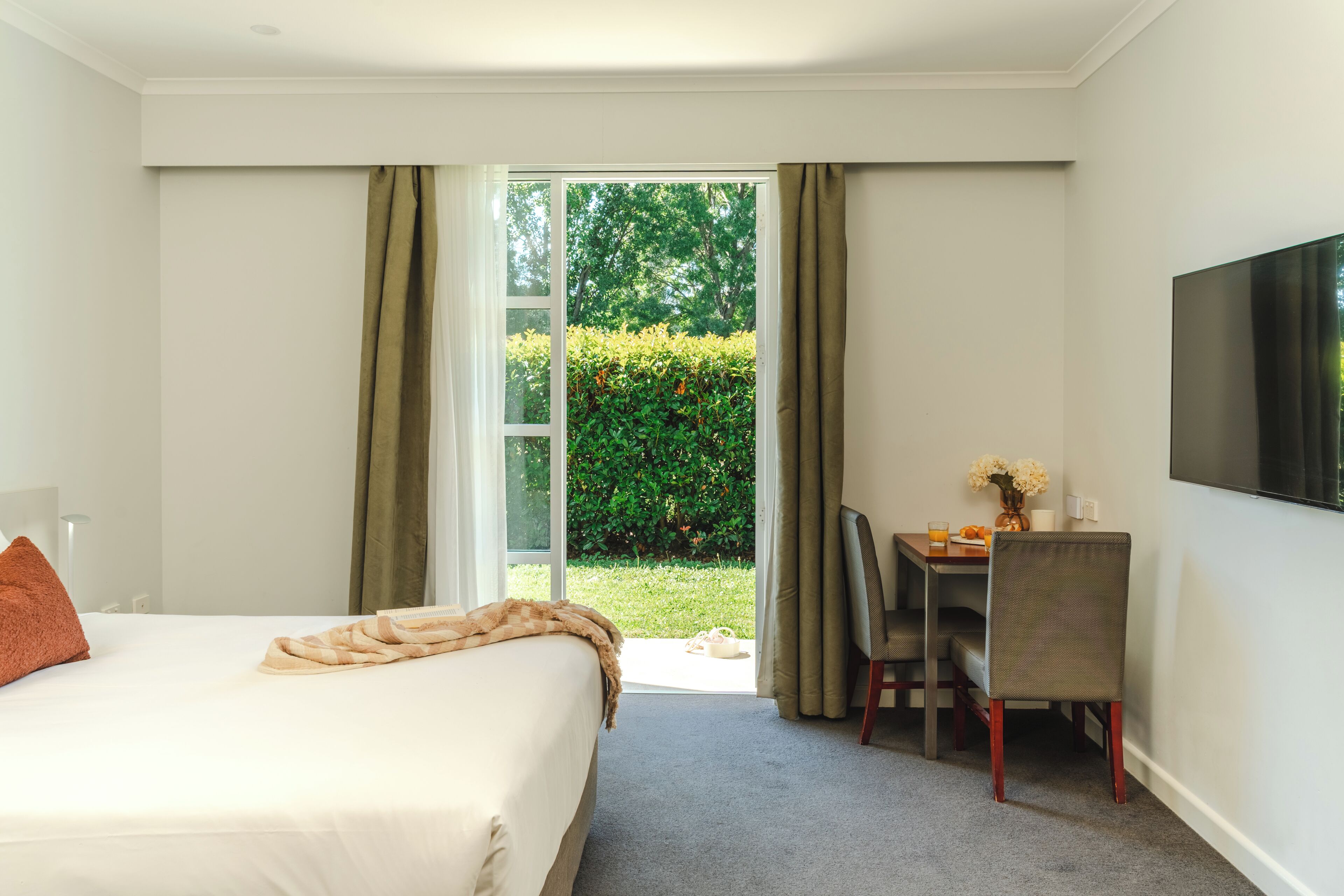 A hotel room with a bed, a desk, a chair, a TV, and a sliding glass door leading to a garden.