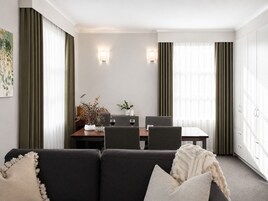 Una habitación de hotel moderna con un sofá de color oscuro, una mesa de comedor con sillas y un amplio ventanal con cortinas.