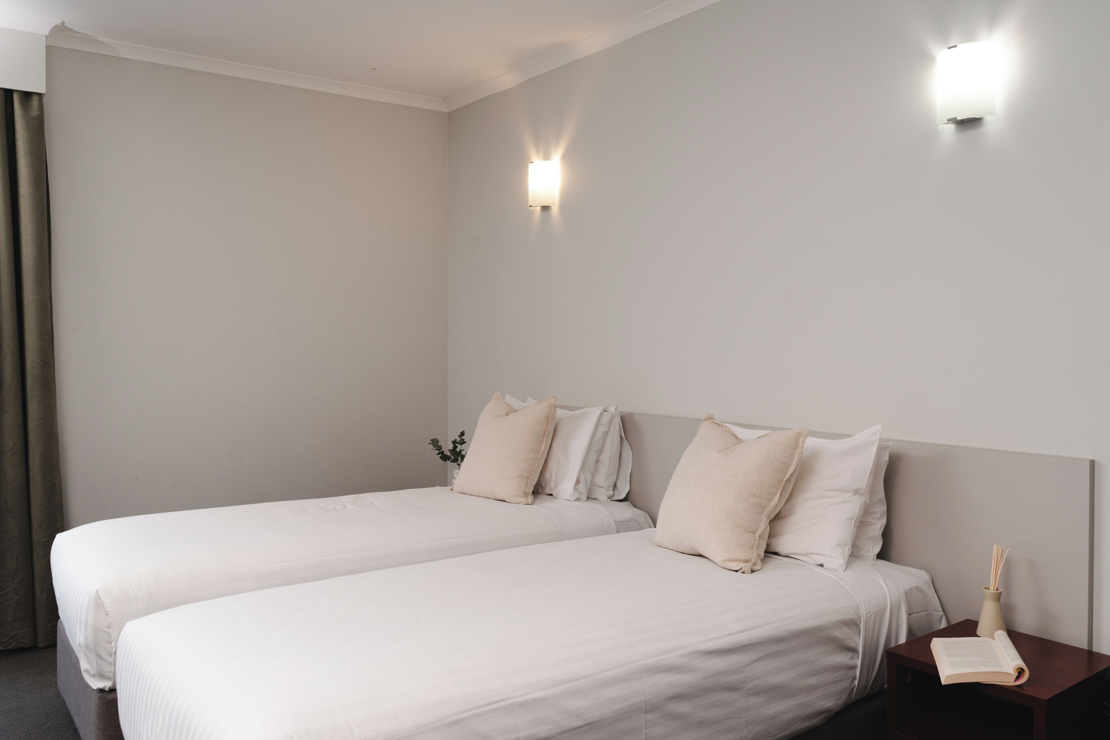 A hotel room with two single beds, a nightstand with a book and a candle, and a wall-mounted light fixture.