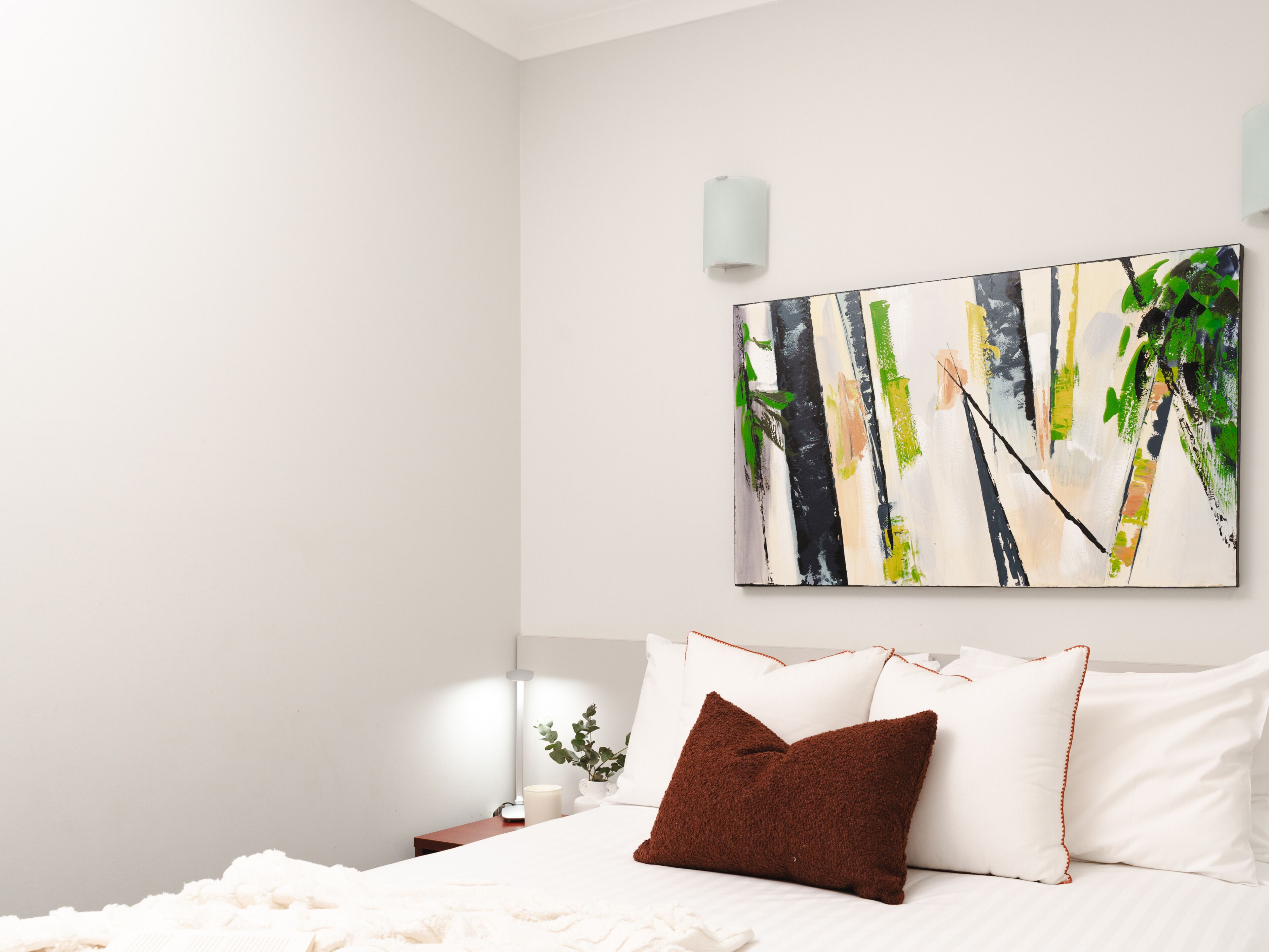 A bedroom with a bed, pillows, a nightstand, a lamp, and a wall-mounted painting.