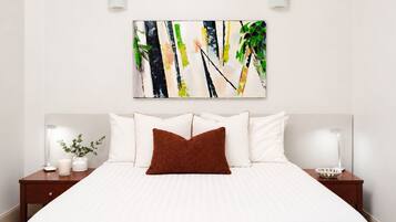 Une chambre d’hôtel avec un lit, du linge de lit blanc, un coussin décoratif marron, deux tables de chevet avec des lampes et un tableau abstrait accroché au mur.