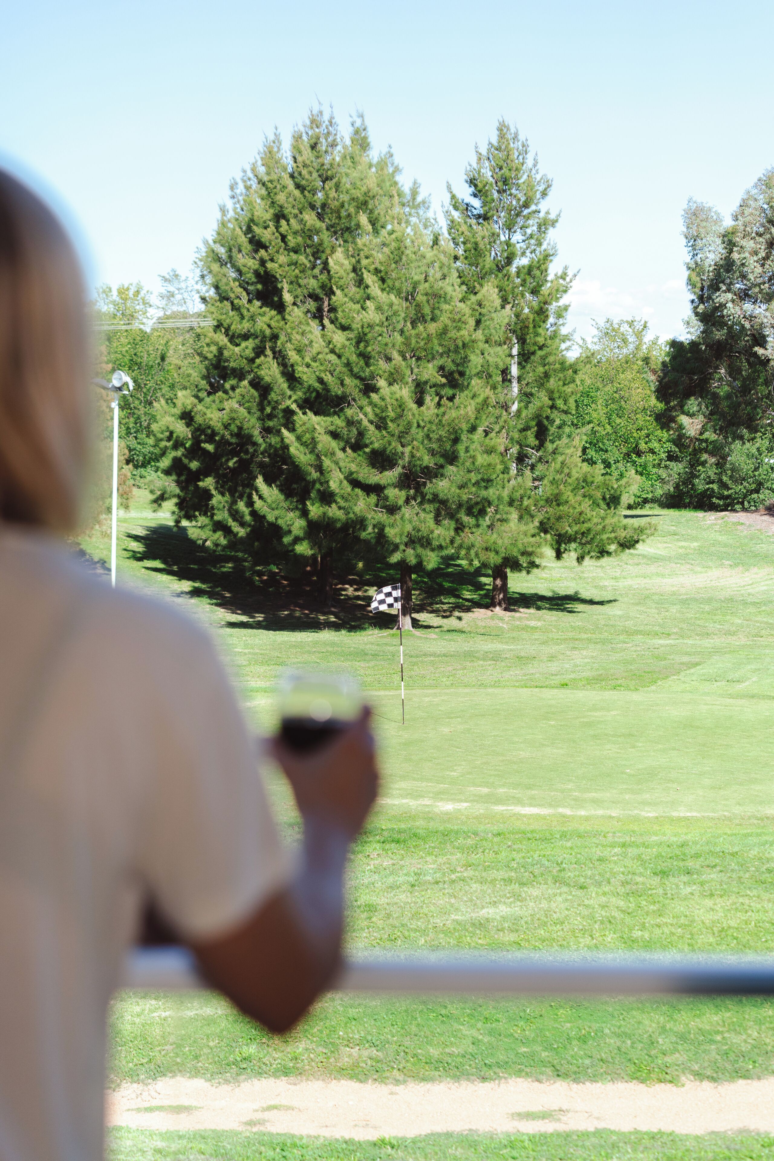 Una persona sosteniendo una bebida, con vista a un campo de golf con árboles y una bandera.