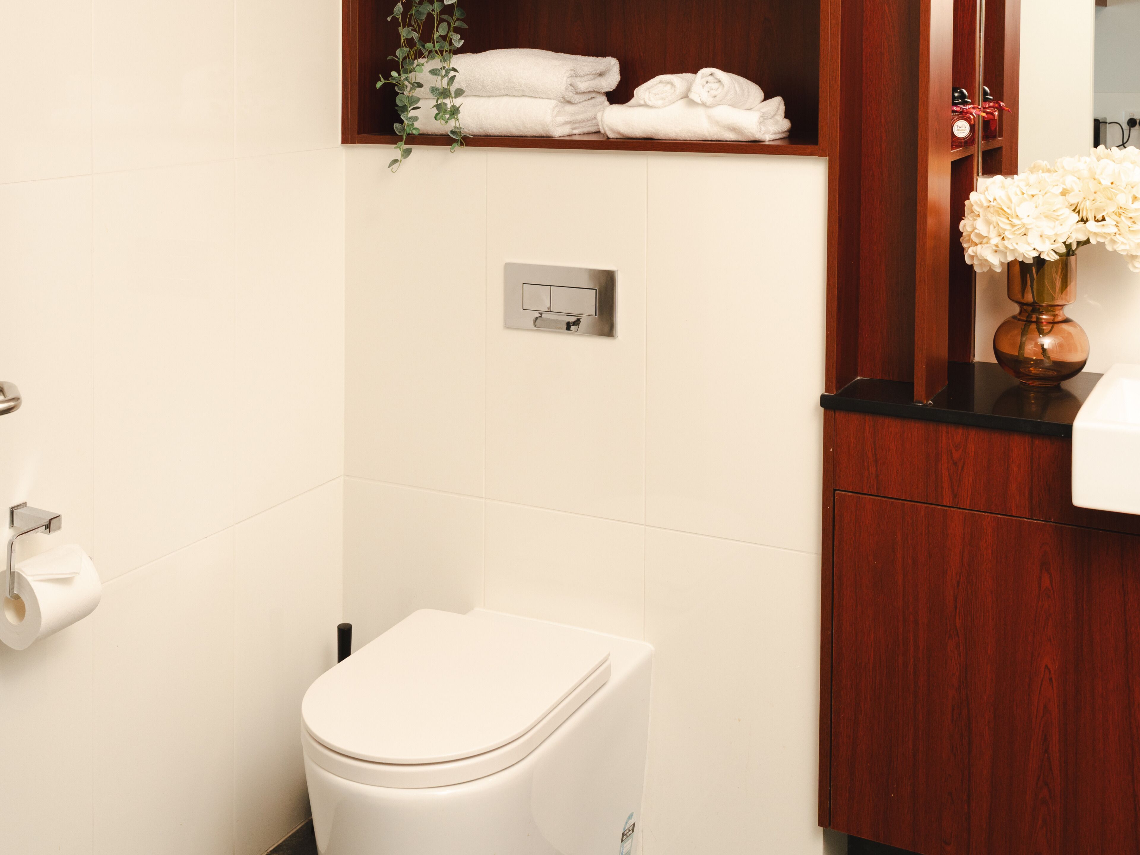 A bathroom with a toilet, a towel rack with folded towels, a wall-mounted electrical outlet, and a wooden cabinet with a vase of flowers.