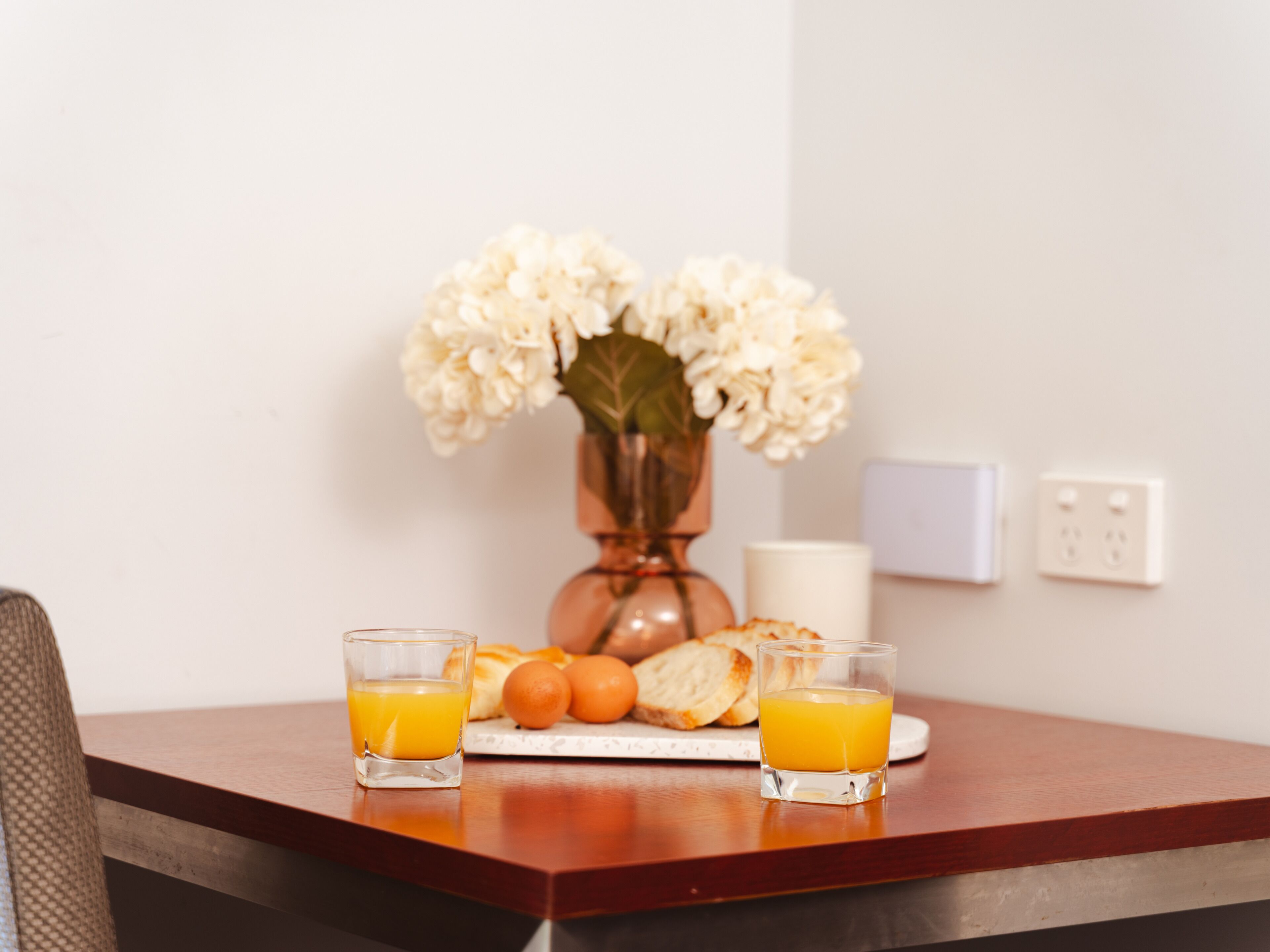 A breakfast table with two glasses of orange juice, a plate of bread, and a vase of flowers.