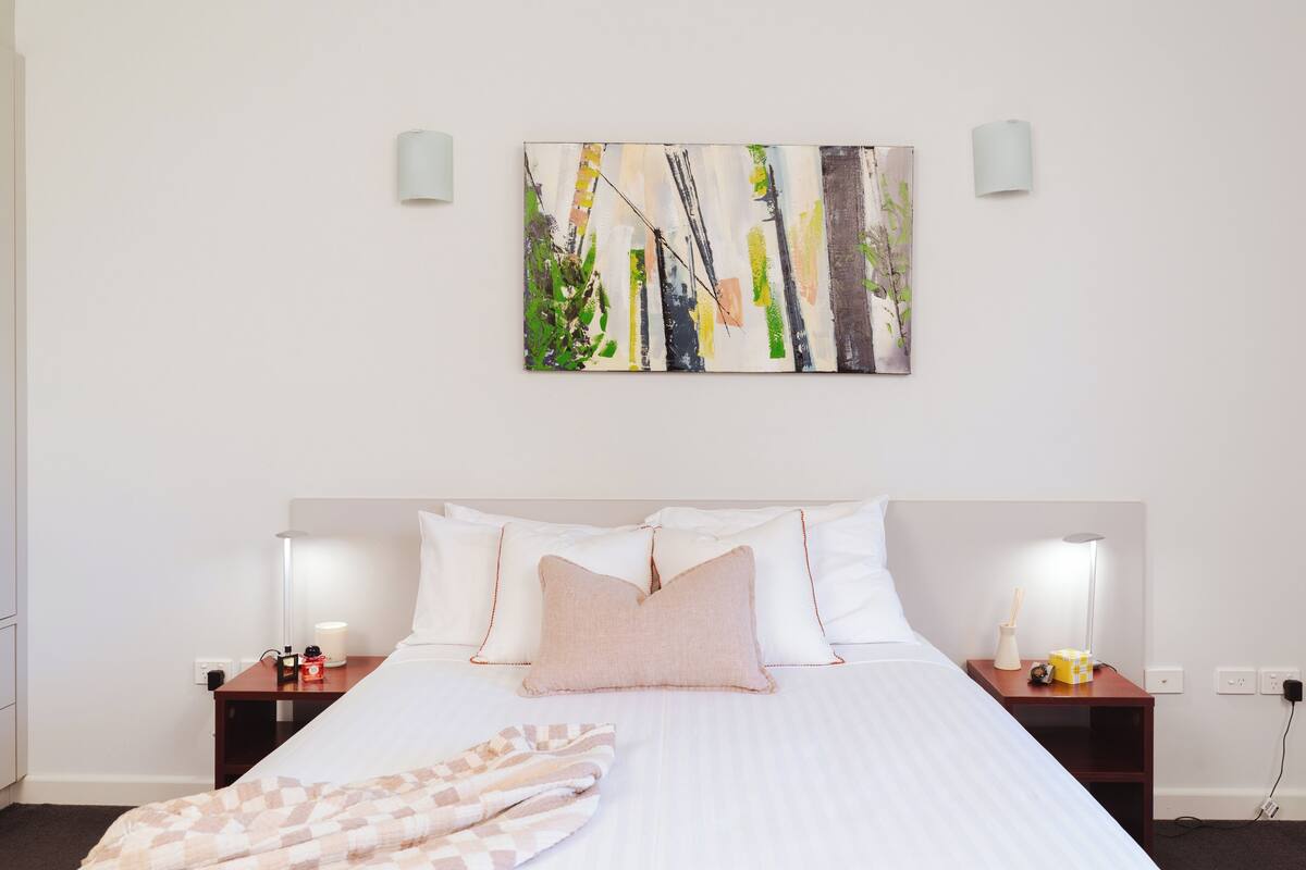 Habitación de hotel con una cama grande, dos mesitas de noche con lámparas, un cuadro colgado en la pared y un enchufe.