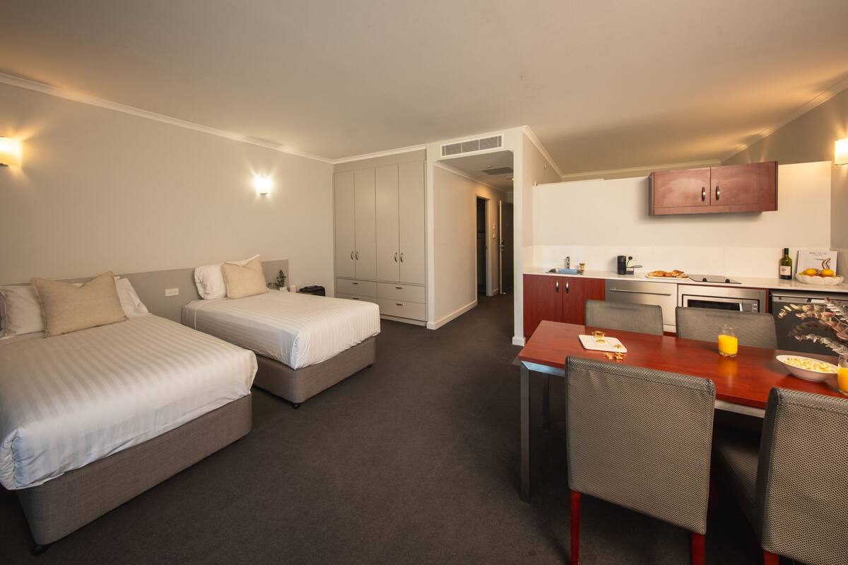 Habitación de hotel con dos camas, una mesa de comedor con sillas, una zona de cocina y un pasillo que conduce a otra habitación.