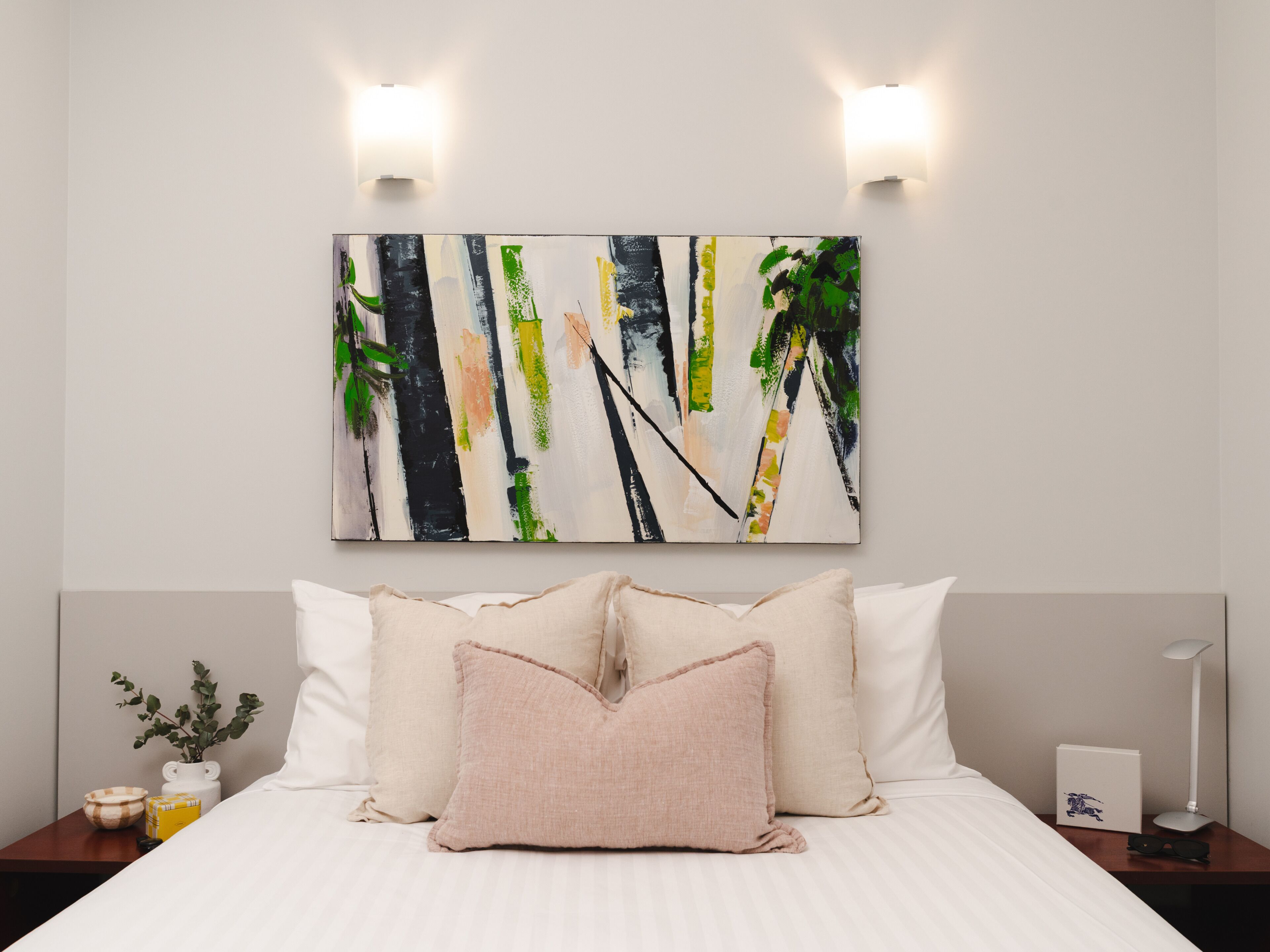A hotel room with a bed, pillows, a nightstand, a lamp, and a wall art piece.