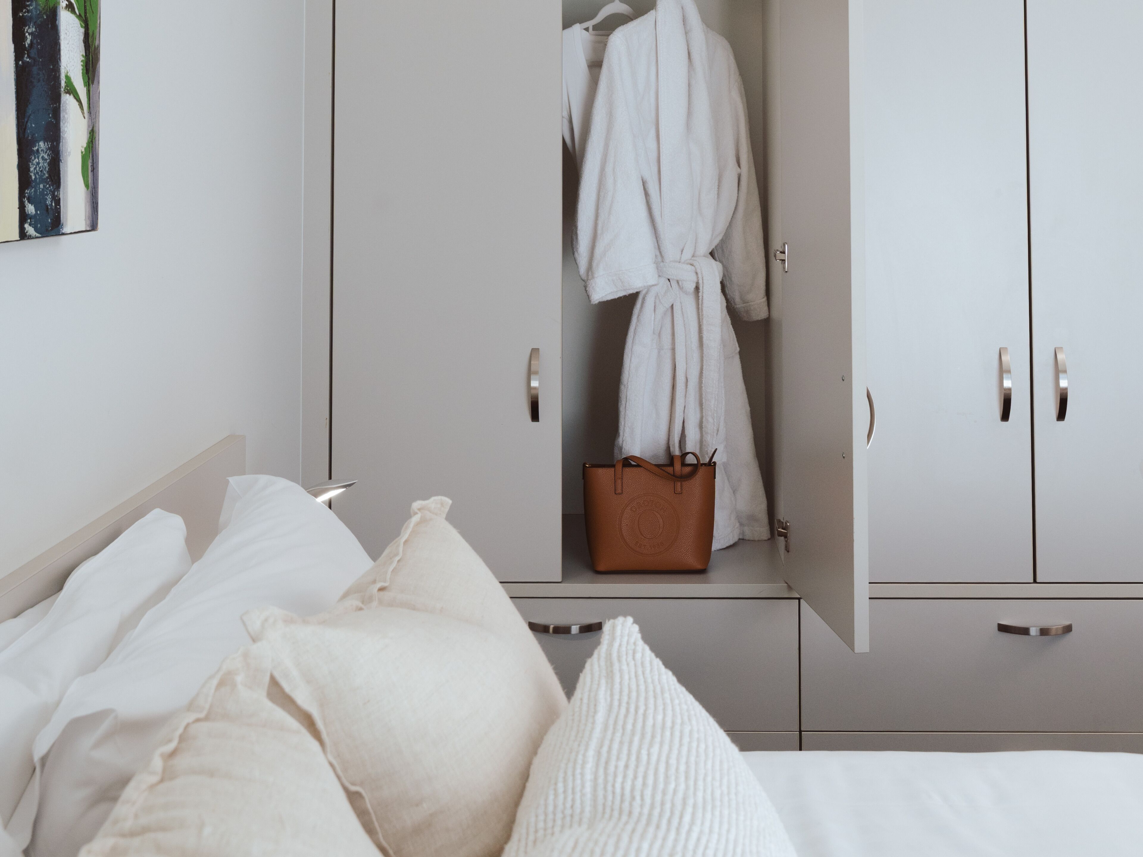 A bedroom with a bed, white pillows, a white robe hanging on a door, and a brown handbag on a shelf.