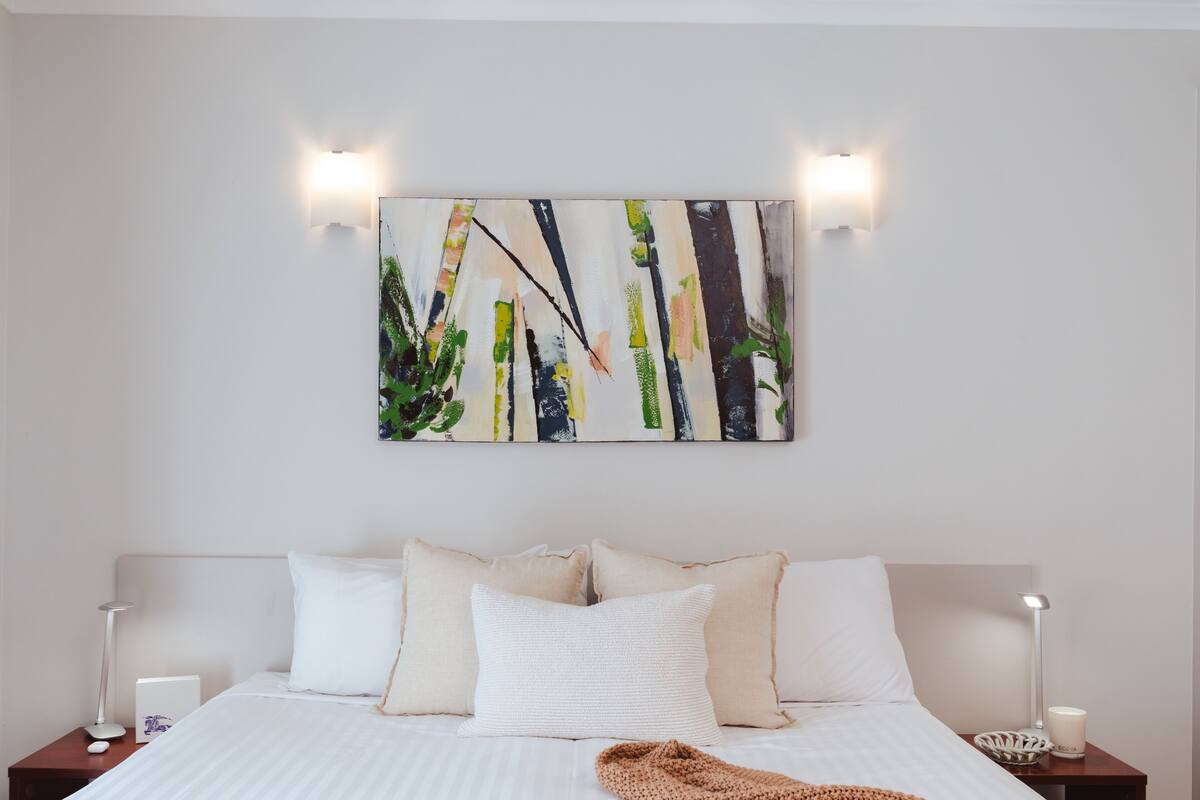 Una cama bien tendida con sábanas blancas, un plaid de punto marrón y un cuadro abstracto moderno encima.