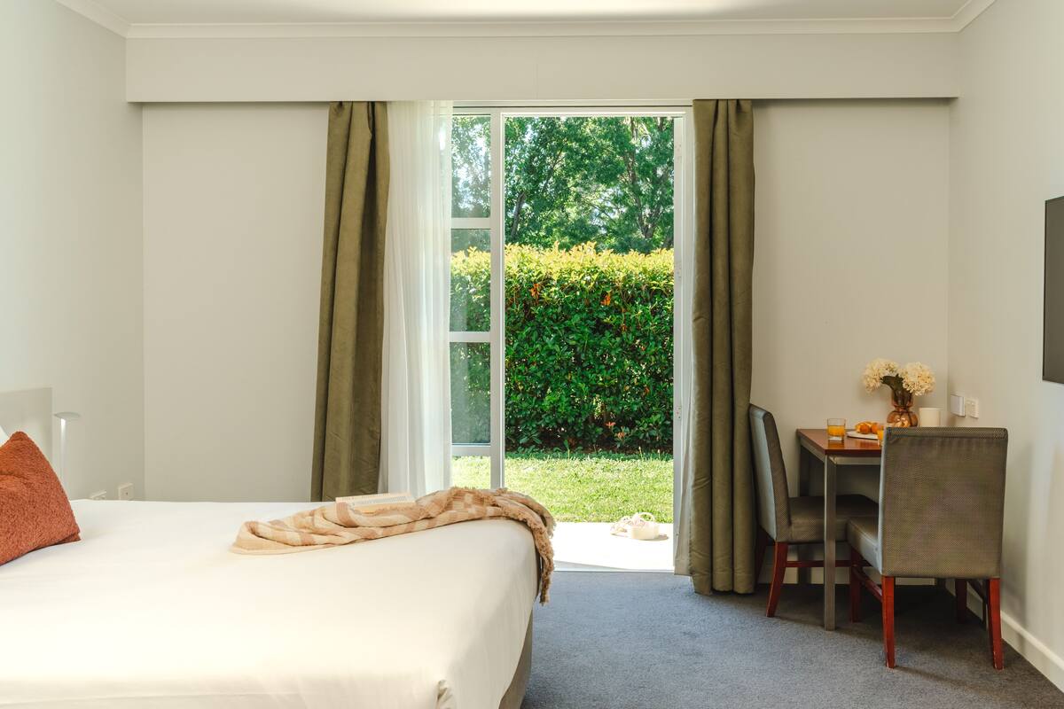 Habitación de hotel con cama, escritorio, sillón y vistas a un jardín a través de una puerta abierta.