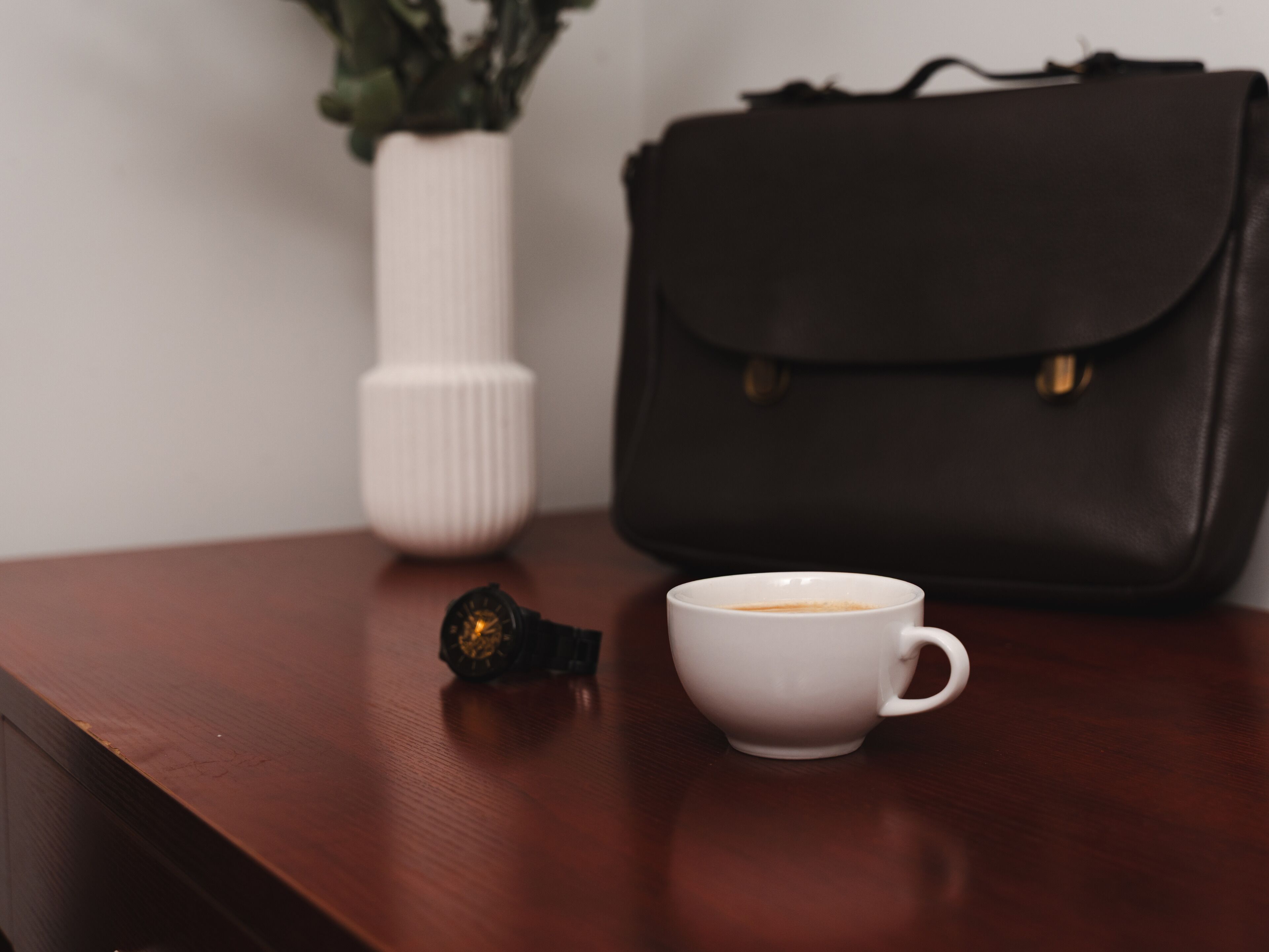 A wooden table with a white cup of coffee, a black bag, and a white vase with a plant.