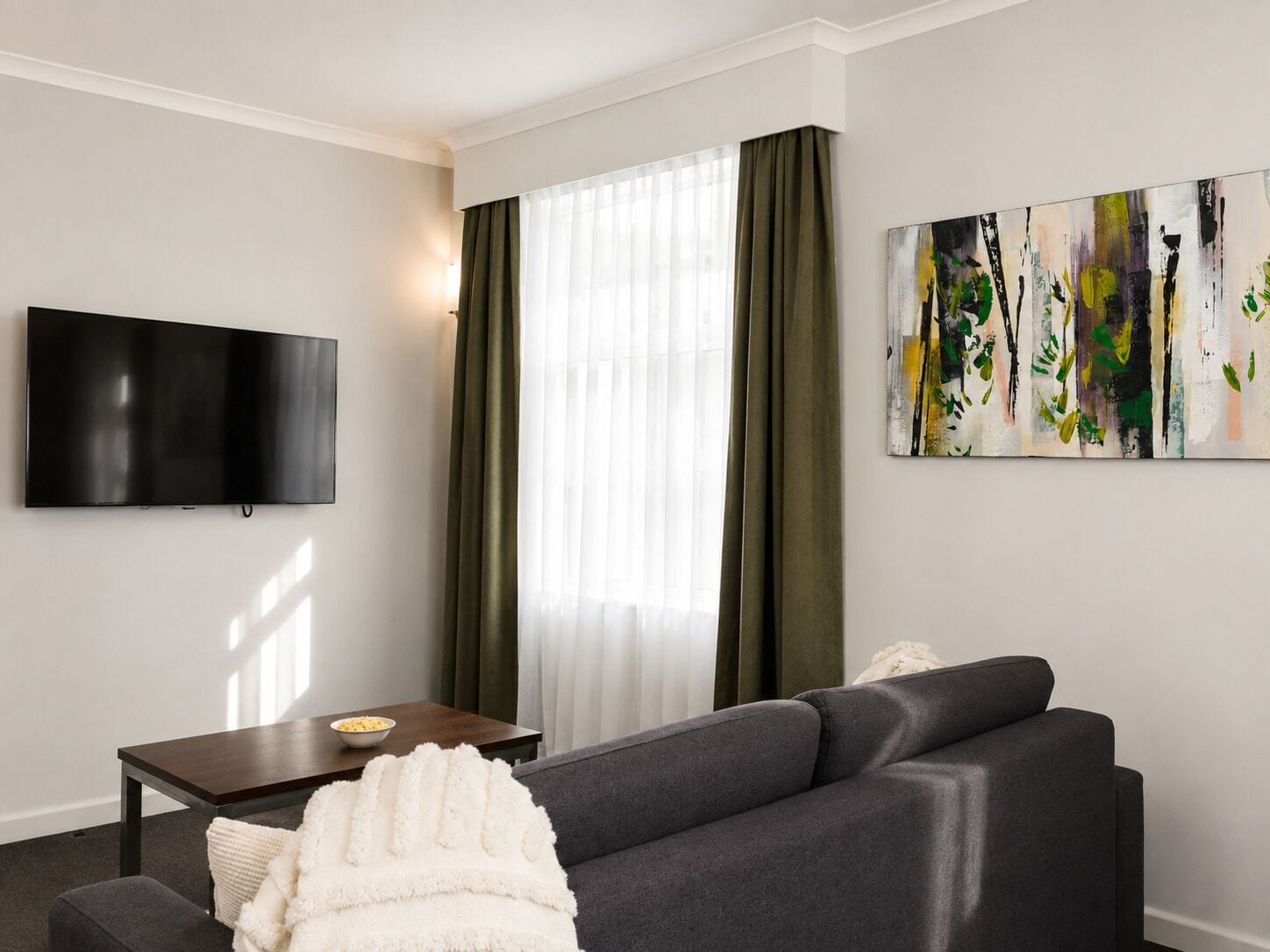 A modern living room with a flat-screen TV, a dark sofa, a small table with a bowl of snacks, and a painting on the wall.