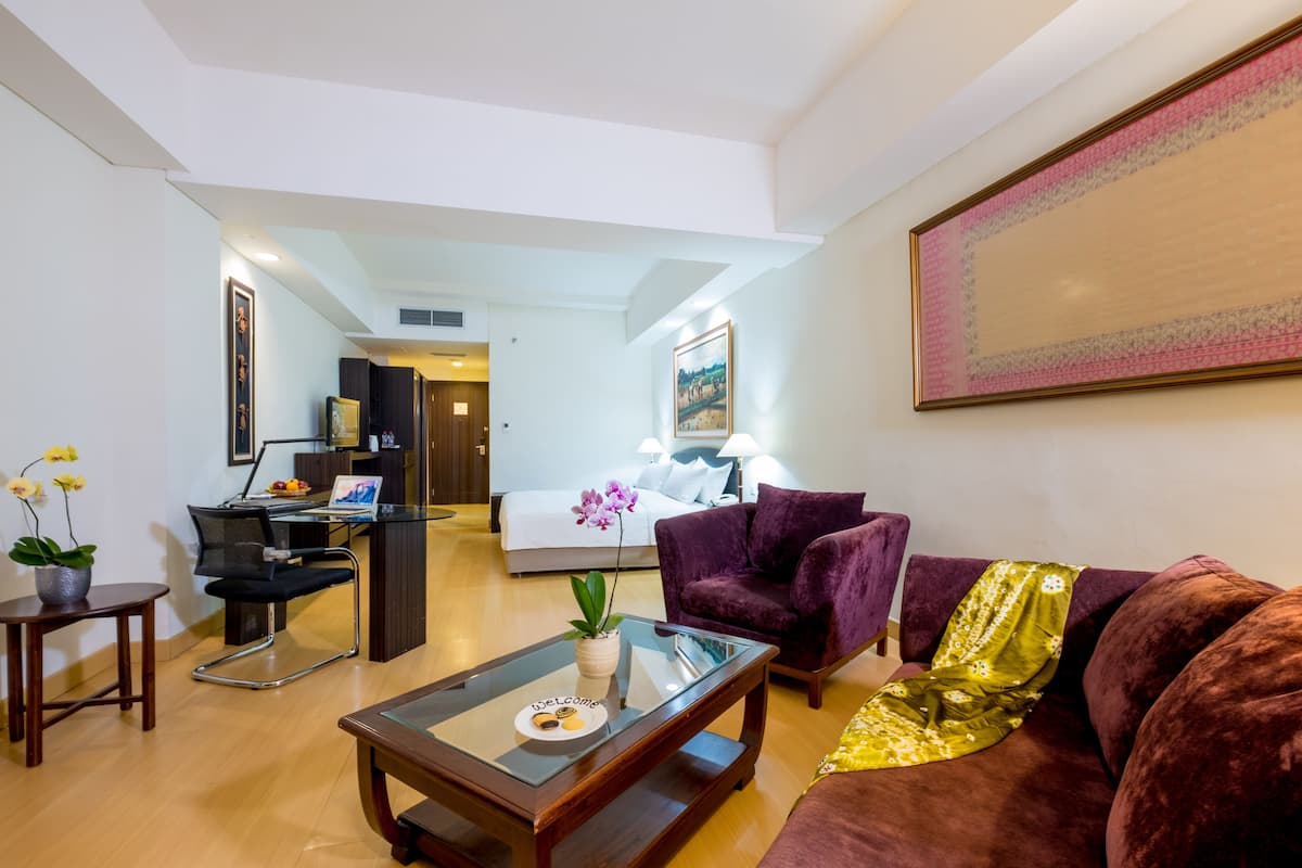Een moderne hotelkamer met een glazen salontafel, een paarse bank, een bruine bank, een bureau met een computer en een bed met een witte dekbedovertrek.