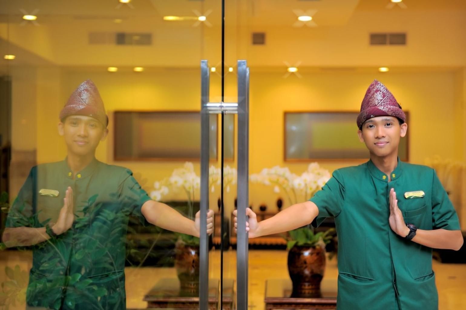 Two individuals in green uniforms standing in front of a glass door with a reflection of a room interior.