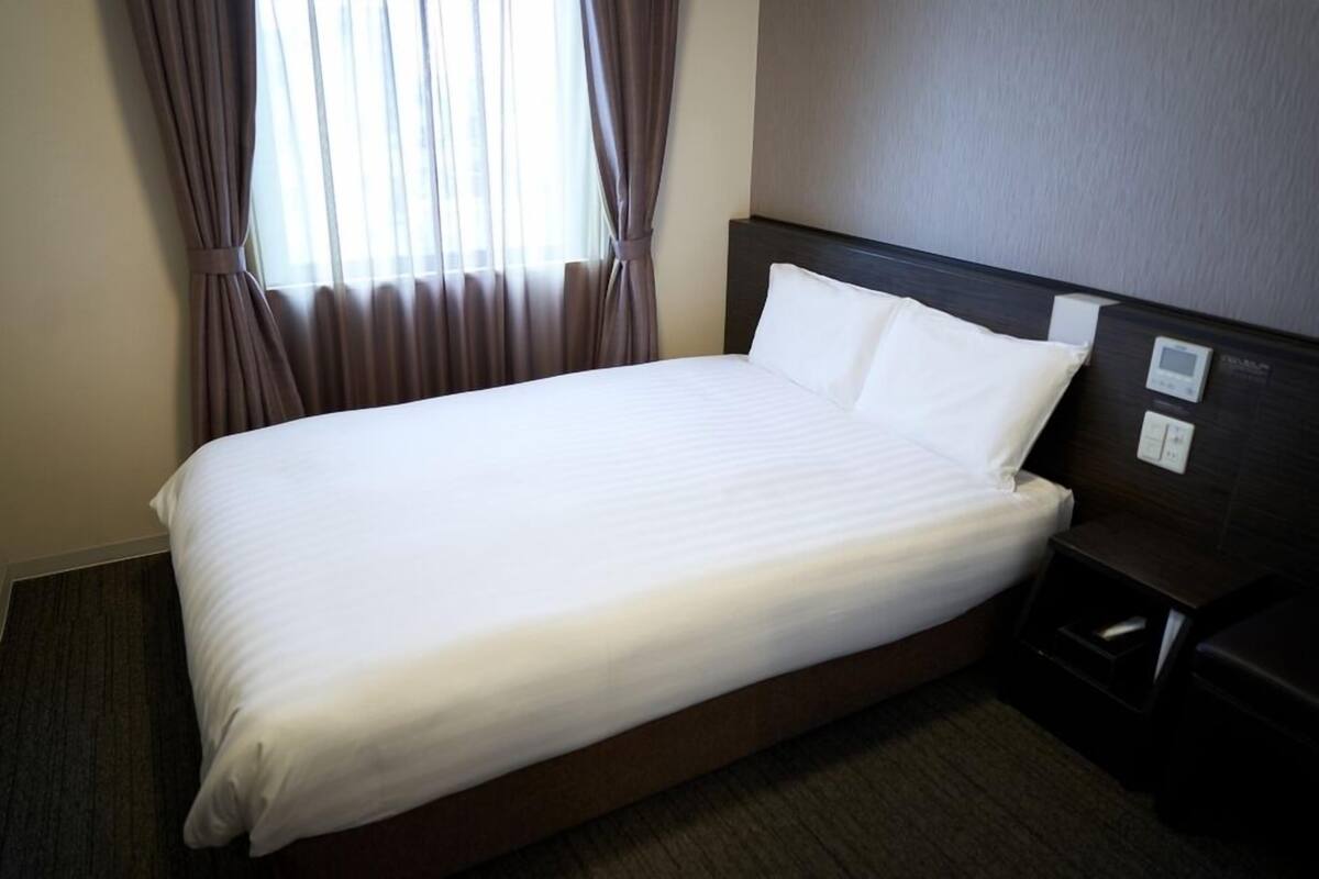 A hotel room with a large bed, a nightstand, a control panel, and a window with curtains.