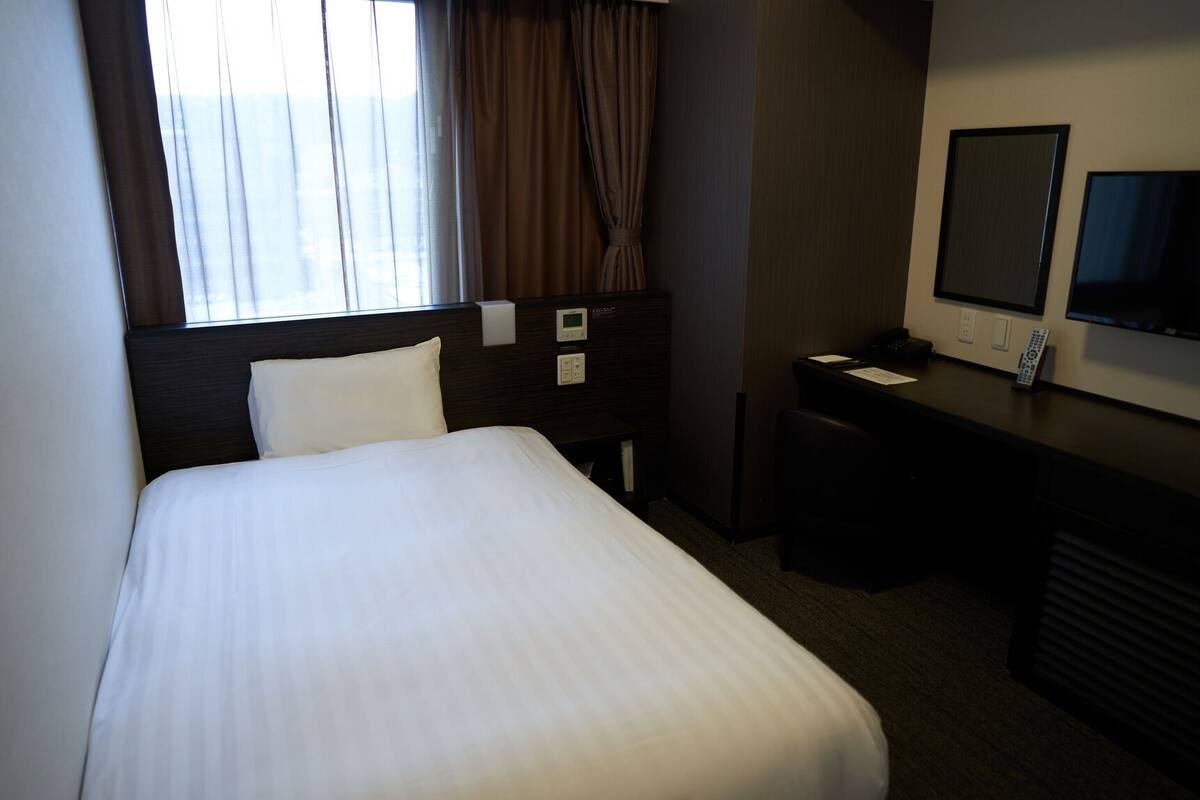 A hotel room with a bed, a desk, a chair, a TV, and a window with curtains.