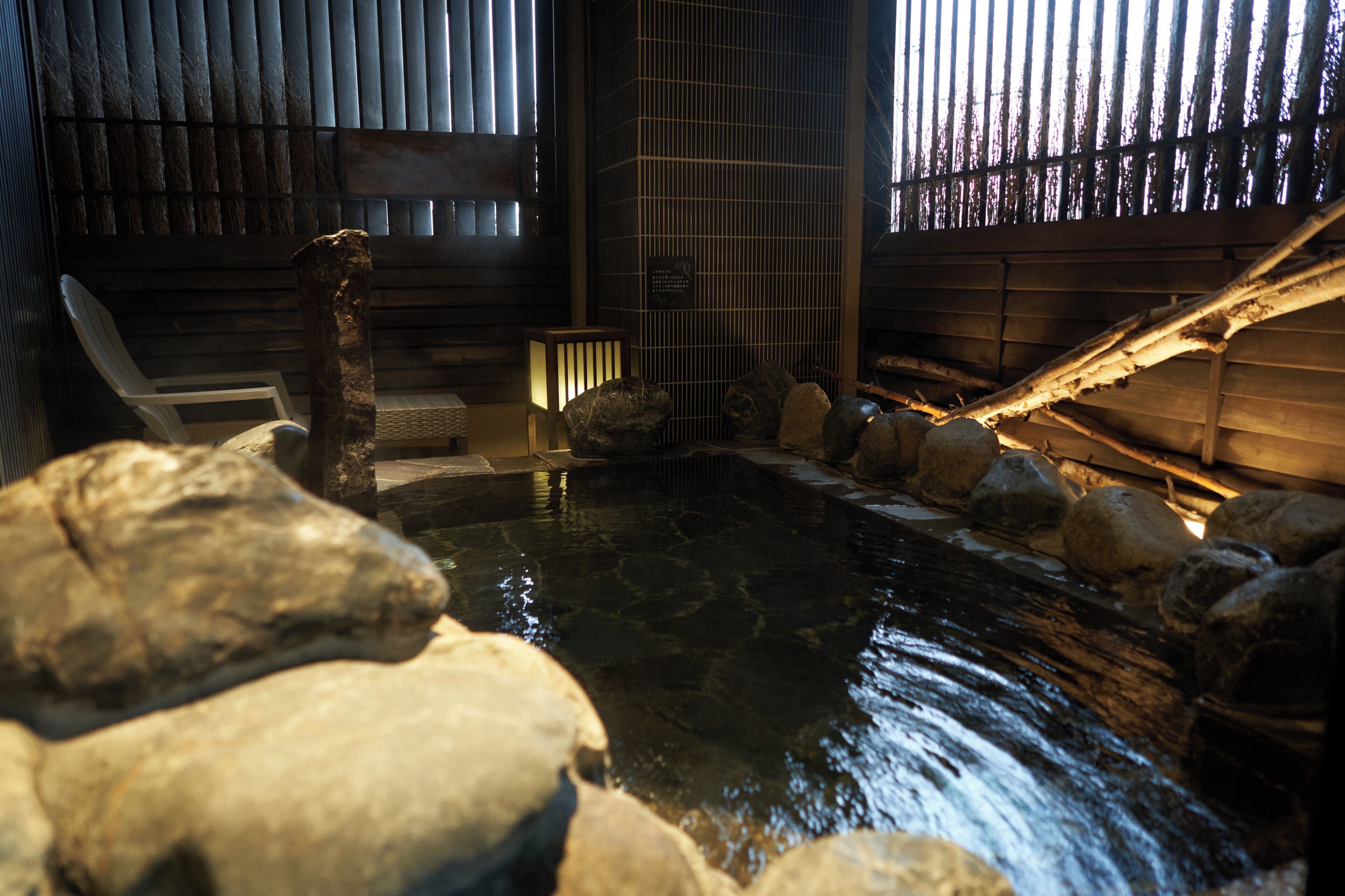 Un onsen japonais traditionnel, avec un intérieur en bois, une source d’eau chaude entourée de pierres et un banc en bois.