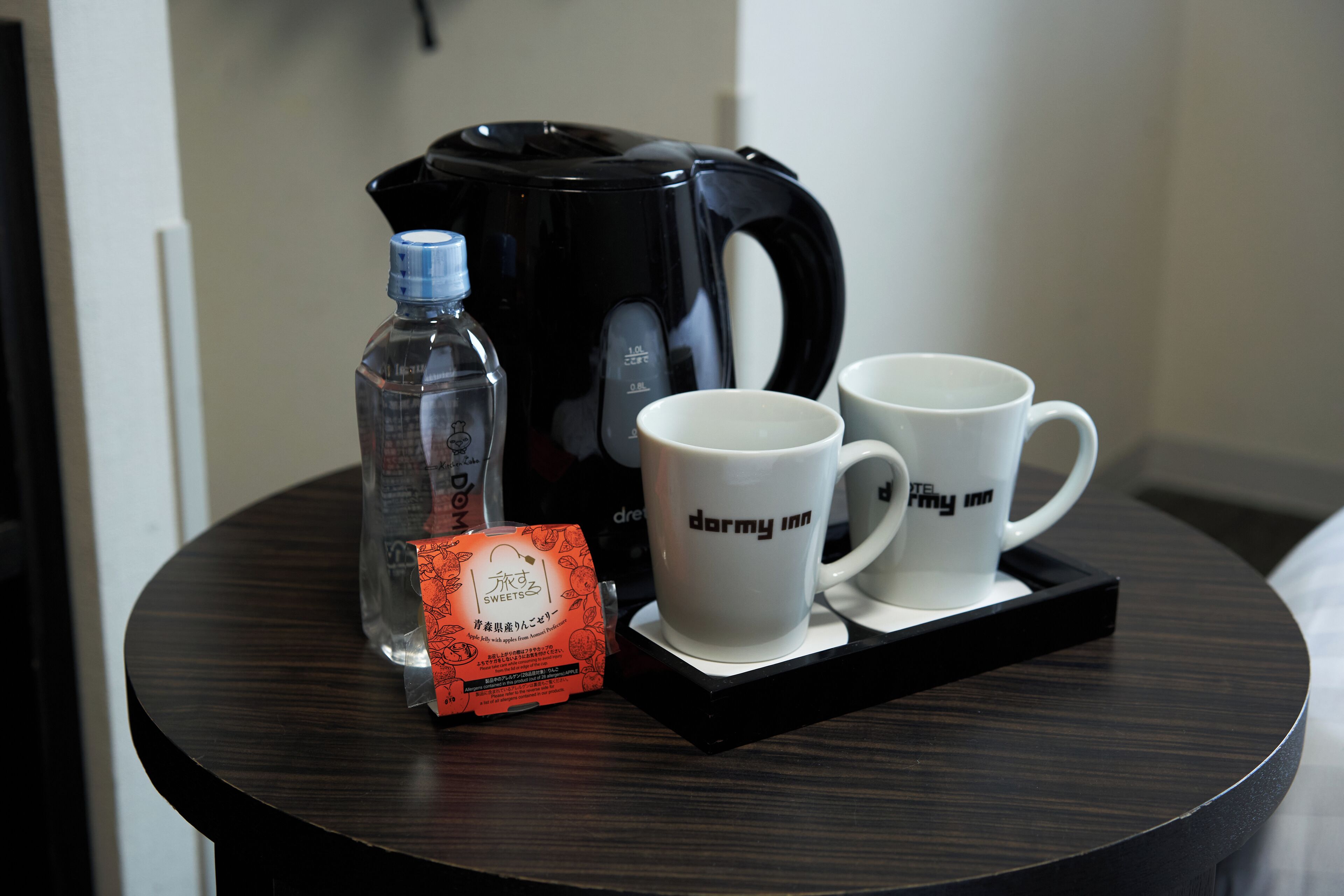 Una mesa con una cafetera negra, dos tazas blancas con el texto "darmy inn", una botella transparente y un paquete de caramelos.