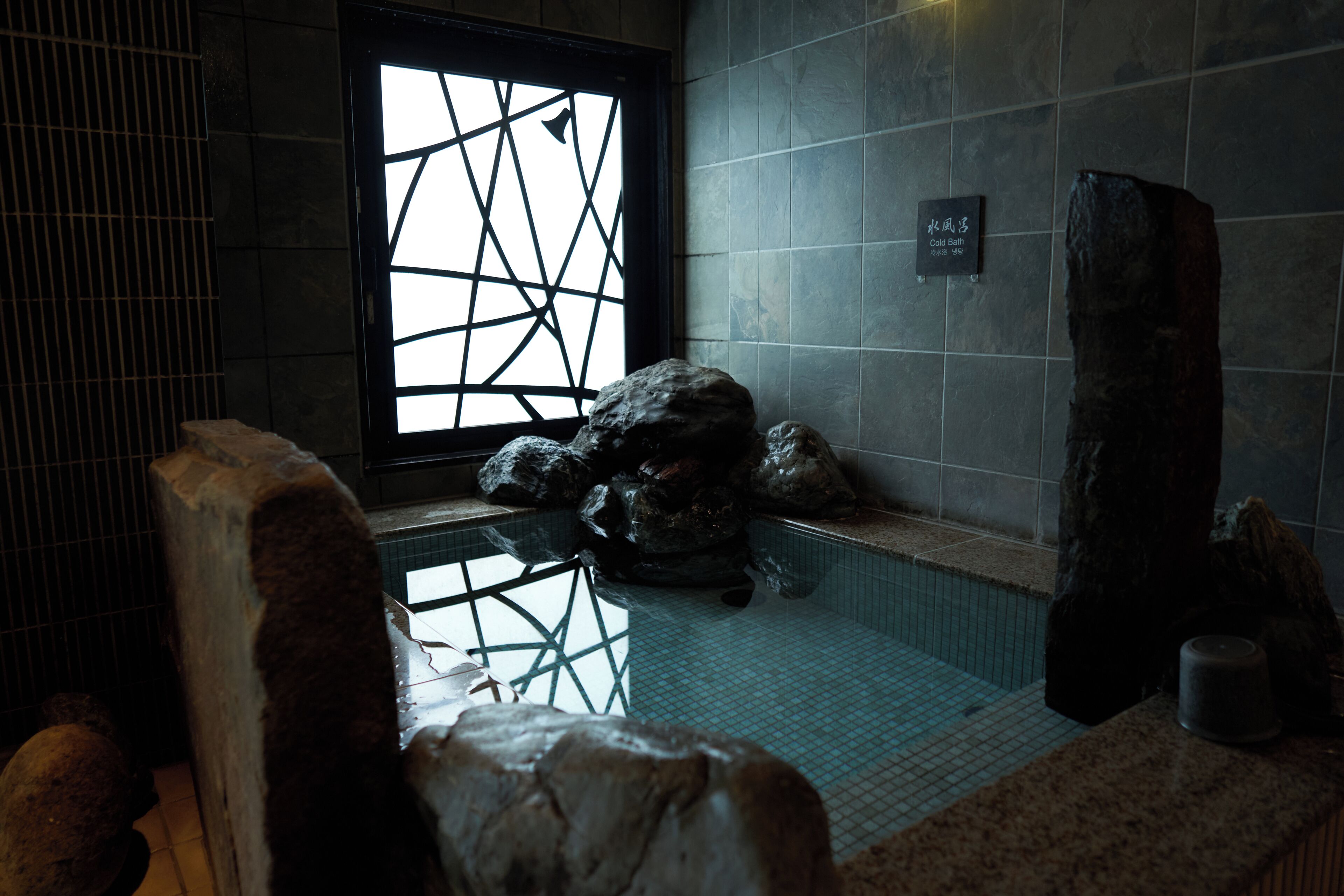 Un área de baño de piedra con suelo de baldosas, un ventanal grande con un diseño geométrico y una pared de piedra con una placa.