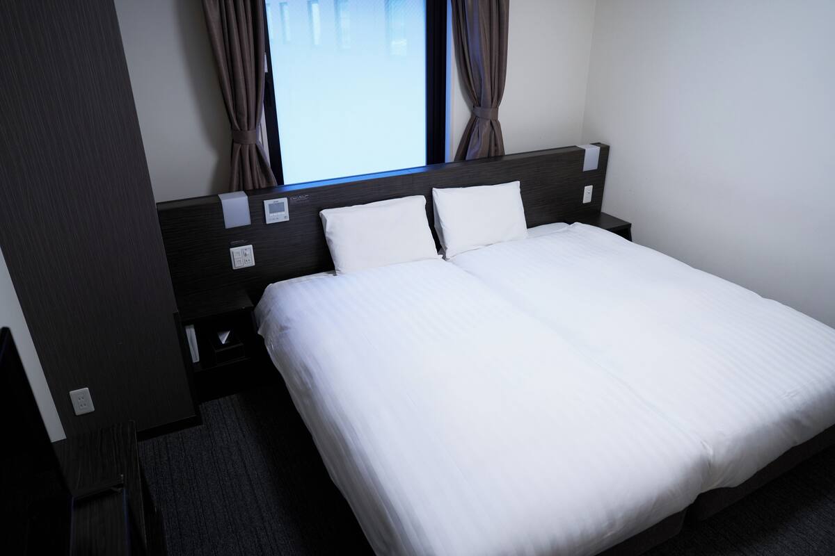 A hotel room with a large bed, two pillows, a headboard, a nightstand, and a window with curtains.