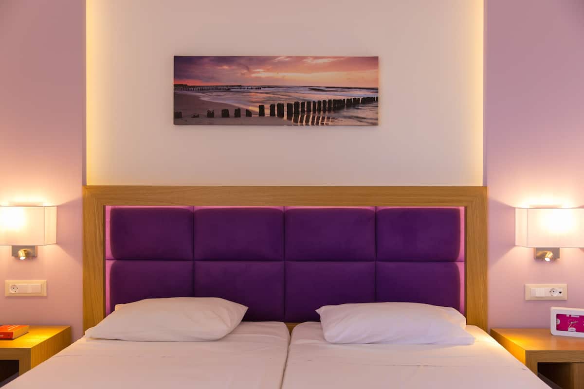 Un lit double avec une tête de lit violette, deux tables de chevet avec des lampes et un tableau accroché au mur représentant un coucher de soleil sur une jetée.