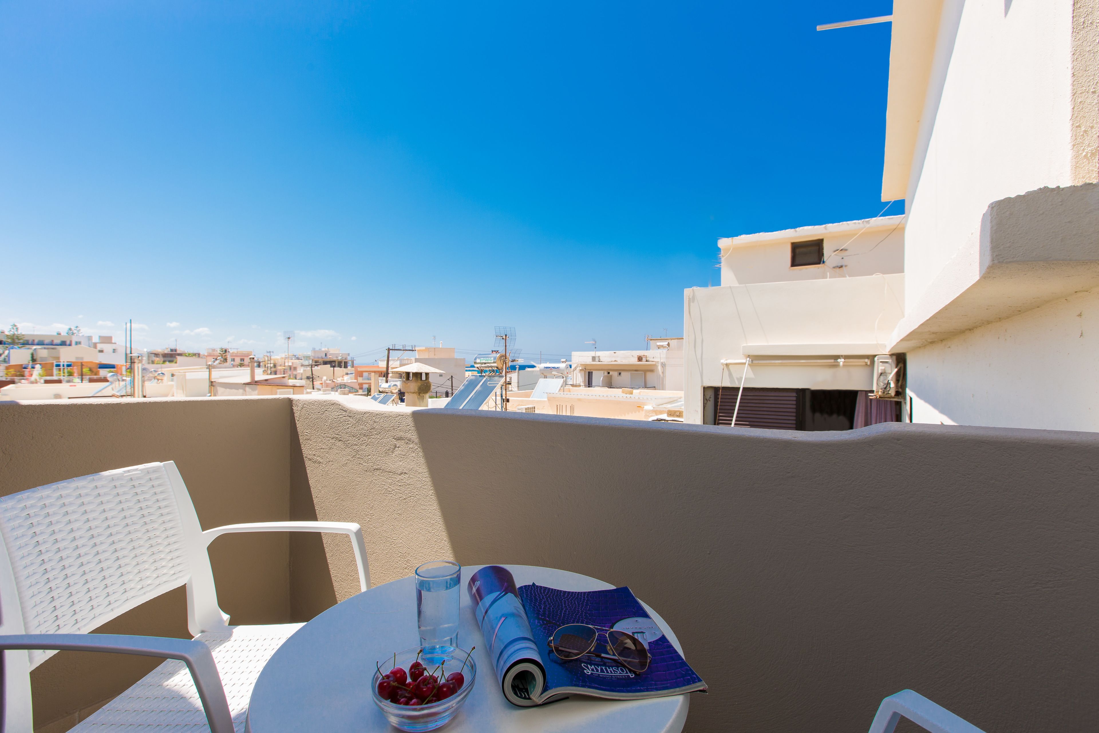 A balcony with a white chair, a table with a magazine, a glass of water, and a bowl of cherries.