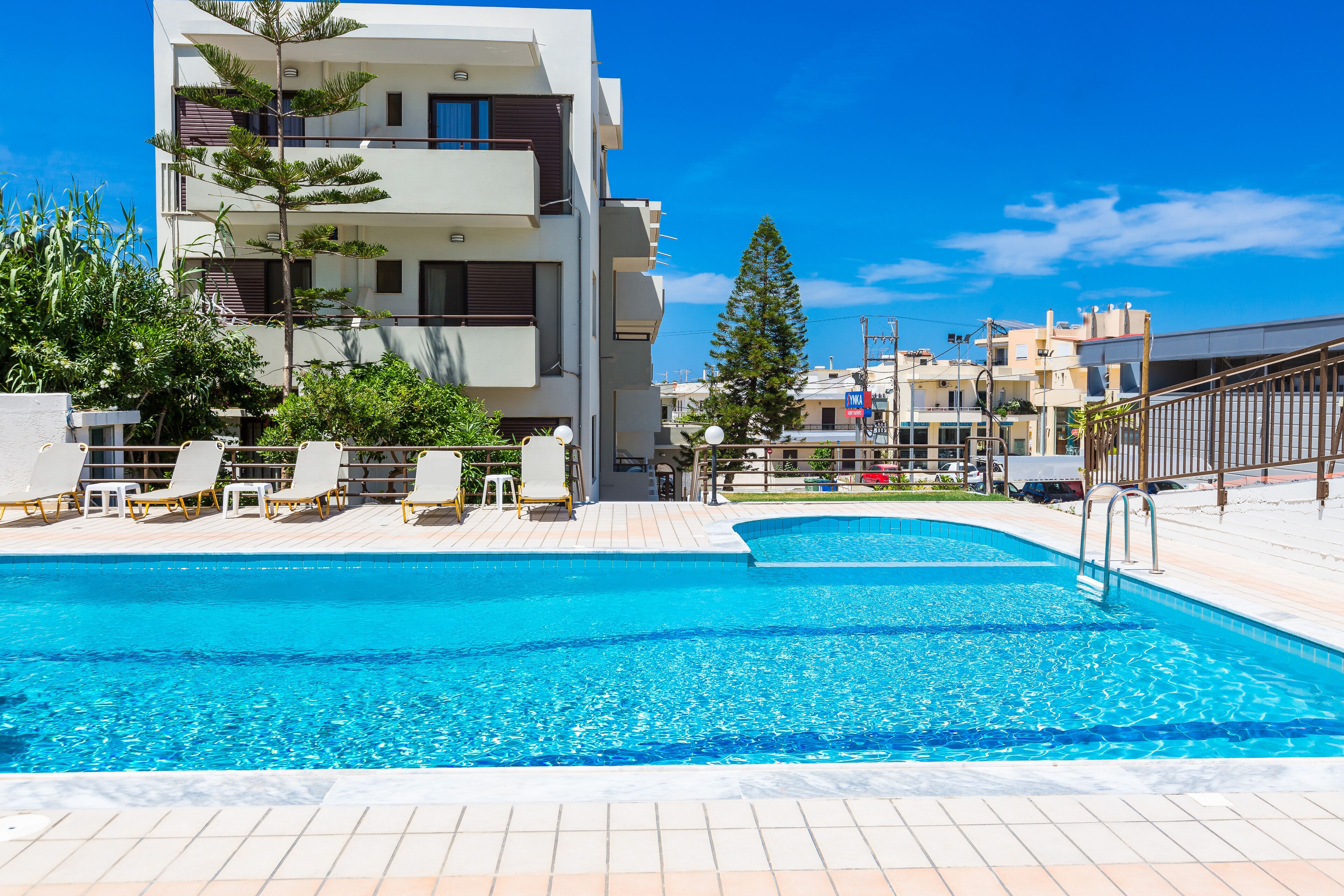 A swimming pool with lounge chairs, a modern building with balconies, and a clear blue sky.
