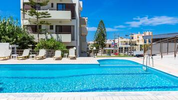 A swimming pool with lounge chairs, a modern building with balconies, and a clear blue sky.