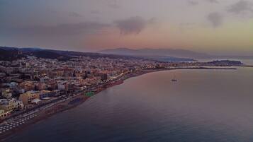 A coastal cityscape with a wide beach, a calm sea, and a distant mountain range.