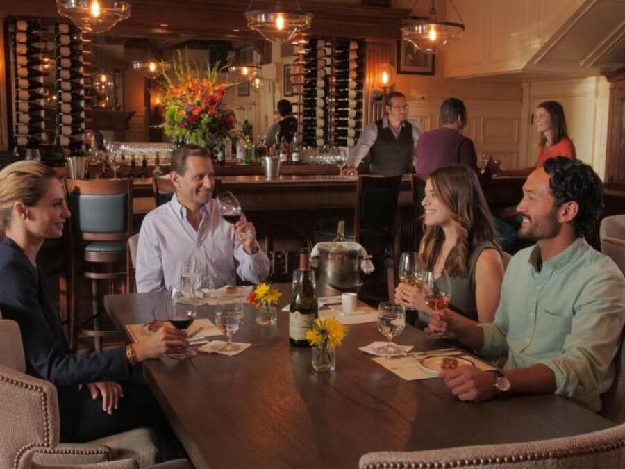 A group of people seated around a table in a bar area with a wine rack and bar counter in the background.