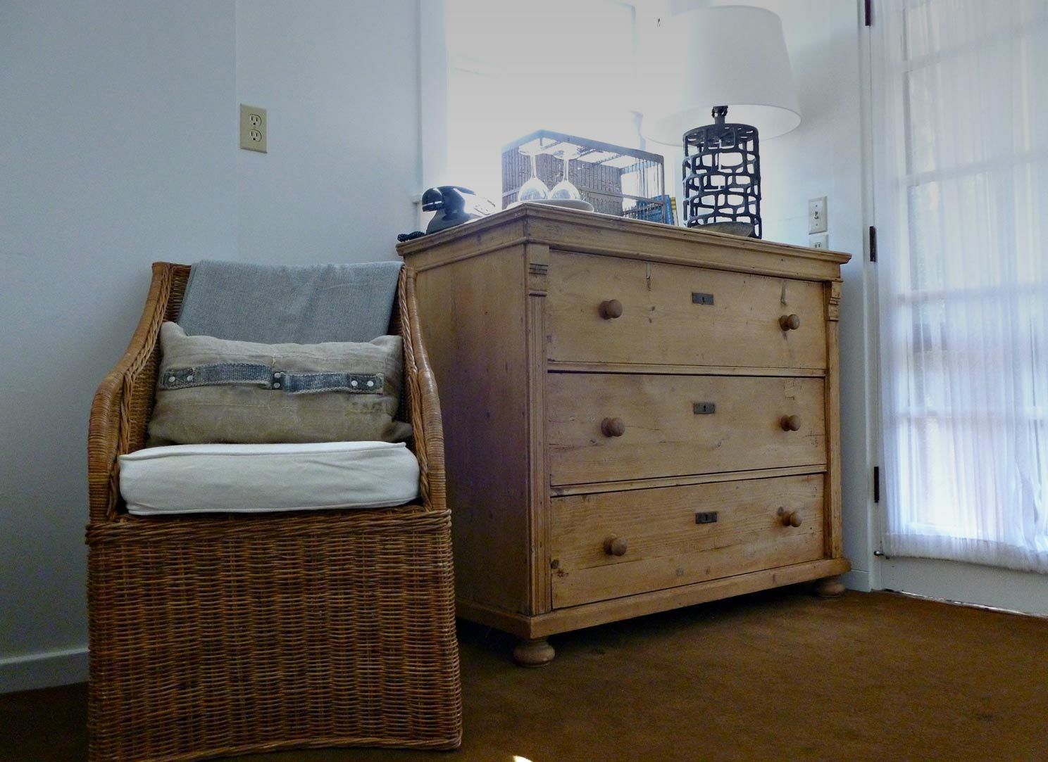 A room with a wicker chair, a wooden chest of drawers, and a lamp.