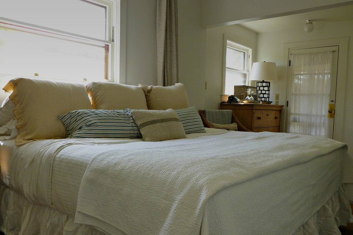 Una cama bien hecha con muchos almohadones y una falda decorativa, una mesita de noche con una lámpara, y una puerta con cortina.