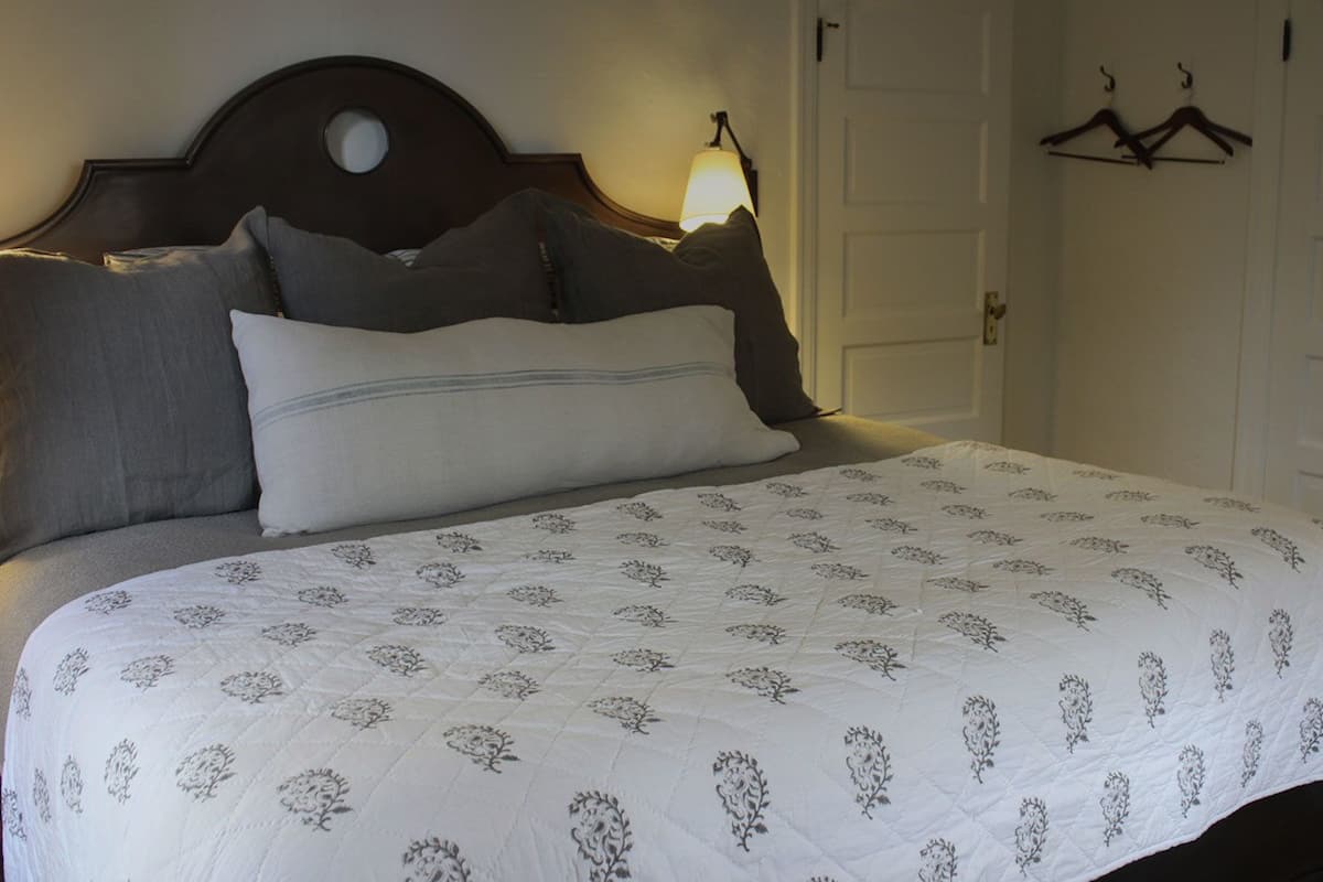 Una cama bien hecha con una colcha de estampado floral y varios almohadones. Una lámpara de noche está encendida, y hay dos perchas en la pared.