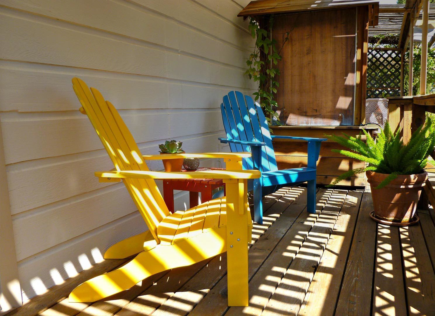 Una terraza de madera con dos sillas Adirondack, una amarilla y otra azul, y una mesita con una planta en maceta.