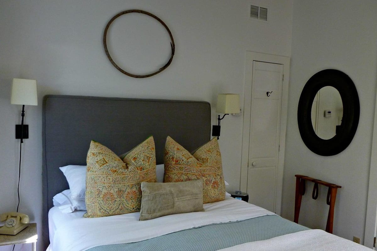 Una cama bien hecha con almohadones estampados, una lámpara de noche, un espejo redondo en la pared y una pequeña silla de madera.