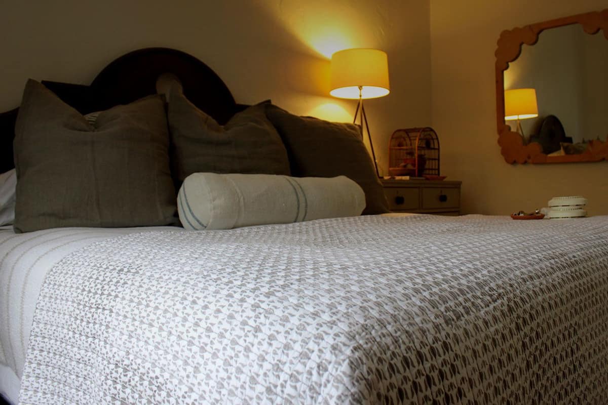 Una cama bien hecha con una colcha estampada, una lámpara de noche, un espejo y una mesita con una lámpara y un objeto decorativo.