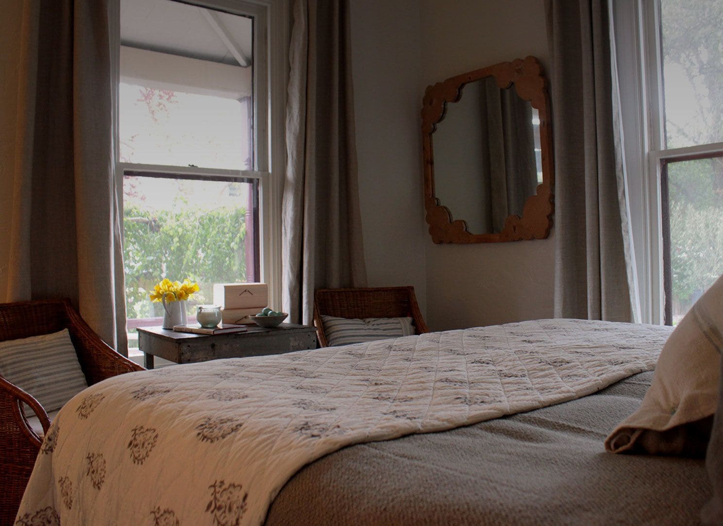 Una cama bien tendida con cubrecama estampado con motivos florales, una mesita de noche de madera con un jarrón de flores amarillas, un espejo en la pared y vistas a la vegetación a través de la ventana.