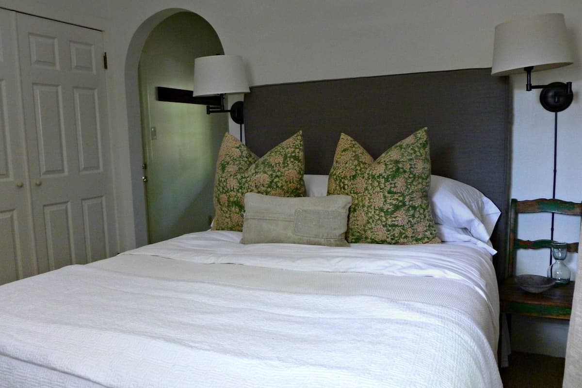 Una cama con sábanas blancas y cuidada presentación, un cabecero oscuro y almohadas con estampado floral. Hay dos lámparas de pared a cada lado de la cama.