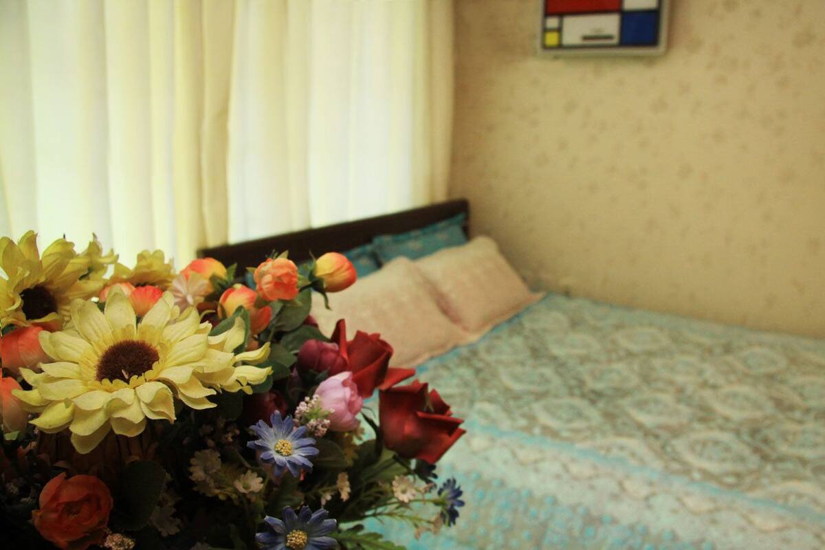 A bed with a patterned bedspread and two pillows, a bouquet of flowers in the foreground, and a wall with a framed picture.