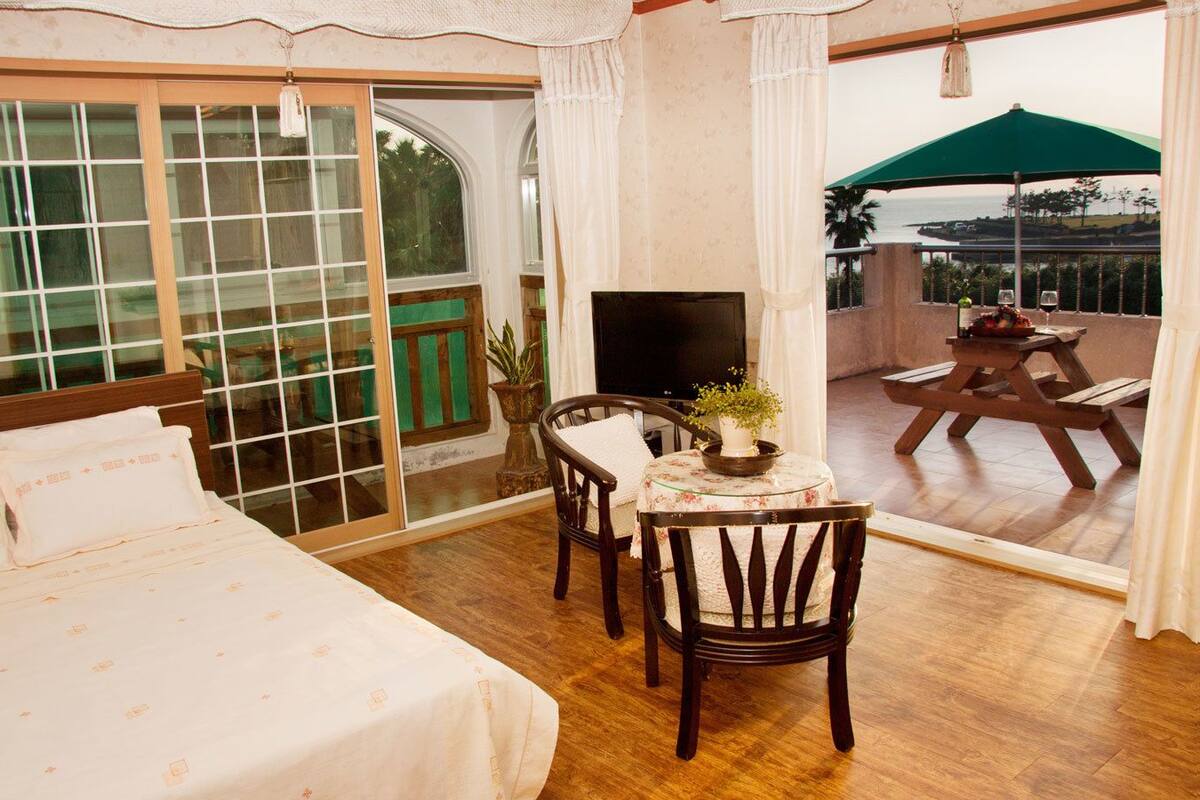 A bedroom with a bed, a TV, a table with chairs, and a view of the outdoors.
