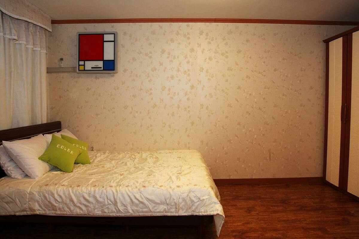 A bedroom with a bed, floral wallpaper, a wooden floor, and a small shelf with a red, white, and blue object.