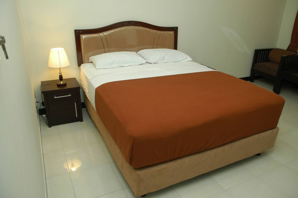 A neatly made bed with a brown bedspread and white pillows, a bedside table with a lamp, and a chair in the corner.