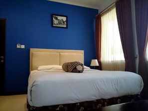 Een hotelkamer met een groot bed, een hoofdbord, een enkele bedlamp, een ingelijst schilderij aan de muur en een raam met gordijnen.