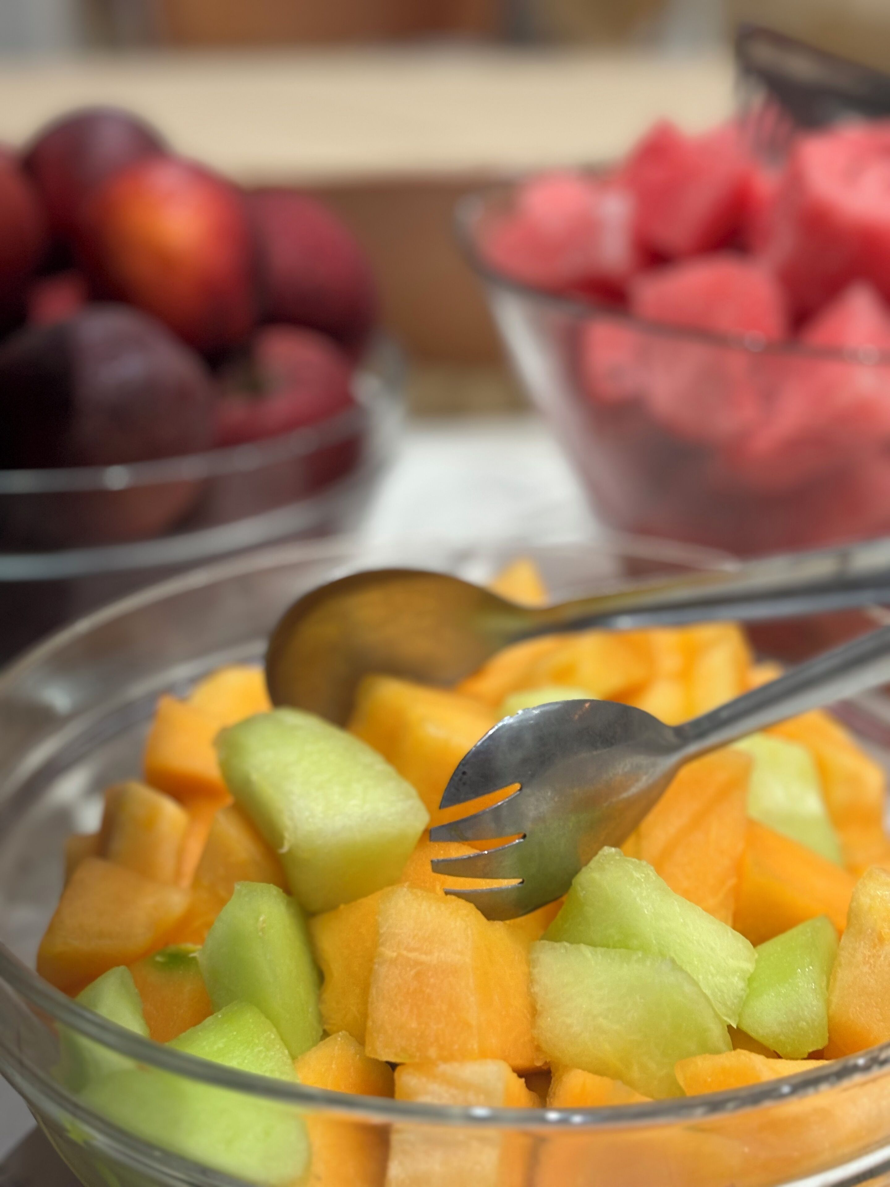 A bowl of cubed cantaloupe with a spoon, with other fruit bowls in the background.