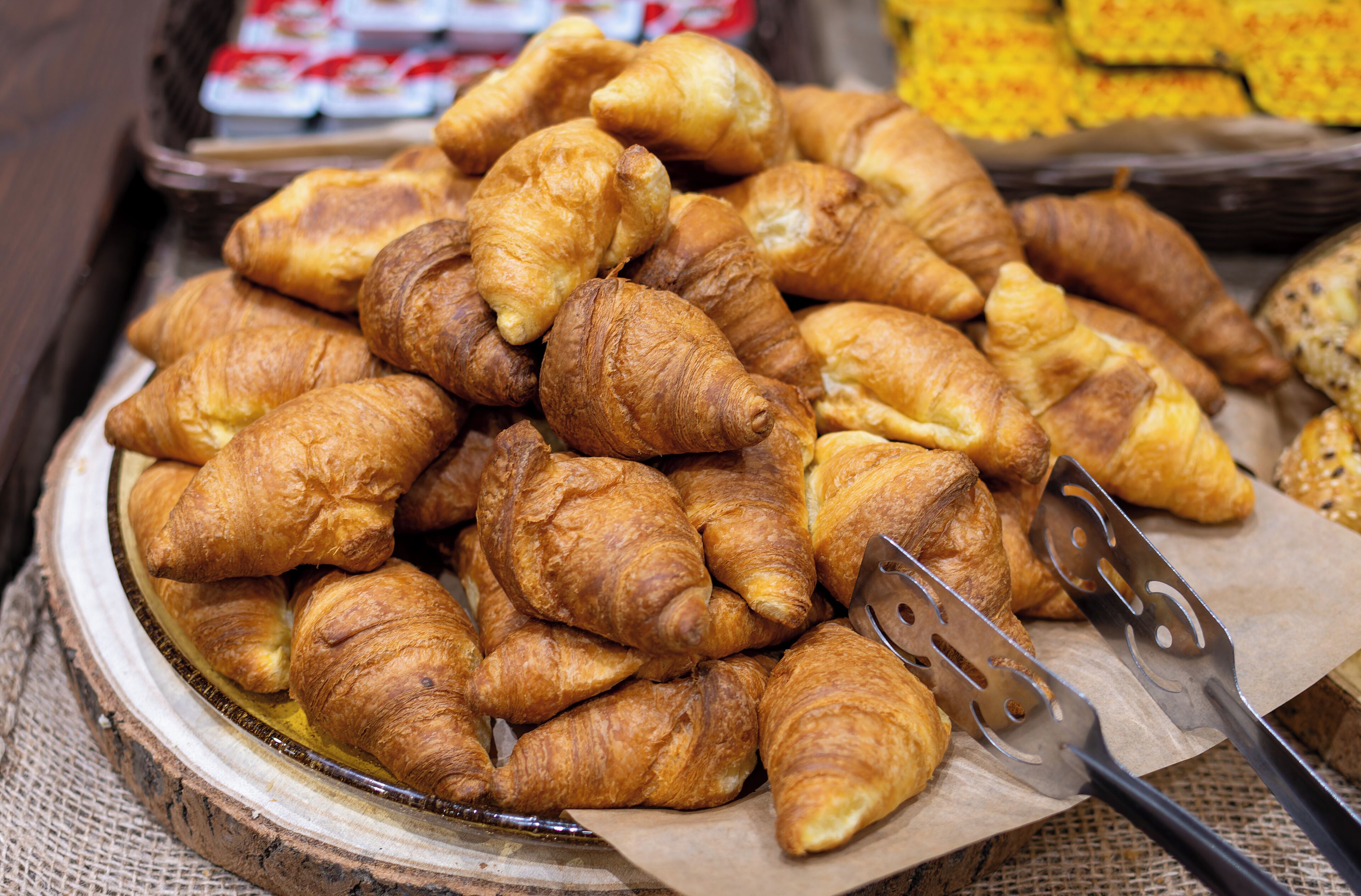 A tray of golden-brown croissants piled on a wooden board, with a pair of tongs resting beside them.